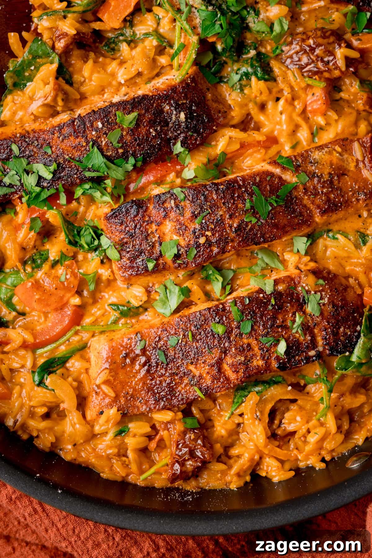 Tuscan Salmon with Creamy Lemon Orzo Recipe 6 Close up overhead image of a dark pan filled with Tuscan-style orzo and pan fried salmon fillets on top, garnished with parsley.