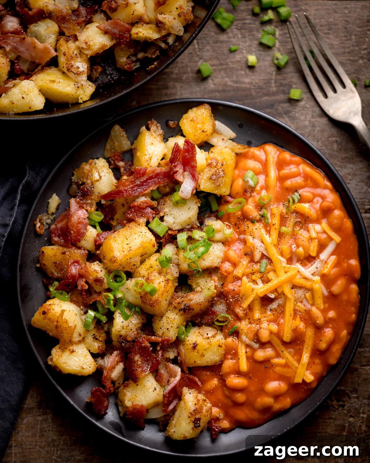 Crispy Potato and Bacon Hash Recipe for Breakfast or Brunch 2 A black plate with potato and bacon hash, roast potatoes, and chopped spring onions on one side, and baked beans topped with grated cheddar cheese on the other. A fork and extra garnish are nearby.