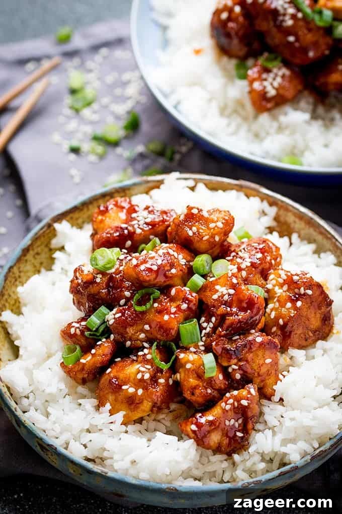Crispy Sesame Chicken with Sticky Sweet-Savory Asian Sauce 3 Portrait photo of Crispy Sesame Chicken with a Sticky Asian Sauce on a bed of rice in a stone bowl with another bowl and chopsticks in the background