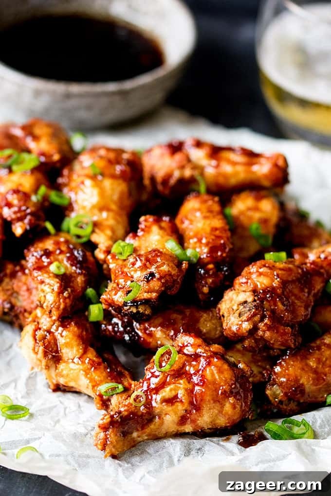 Sticky Crispy Asian-Style Chicken Wings Recipe 3 Portrait photo of Sticky and Crispy Asian Chicken Wings with a bowl of sauce and a glass of beer in the background out of focus