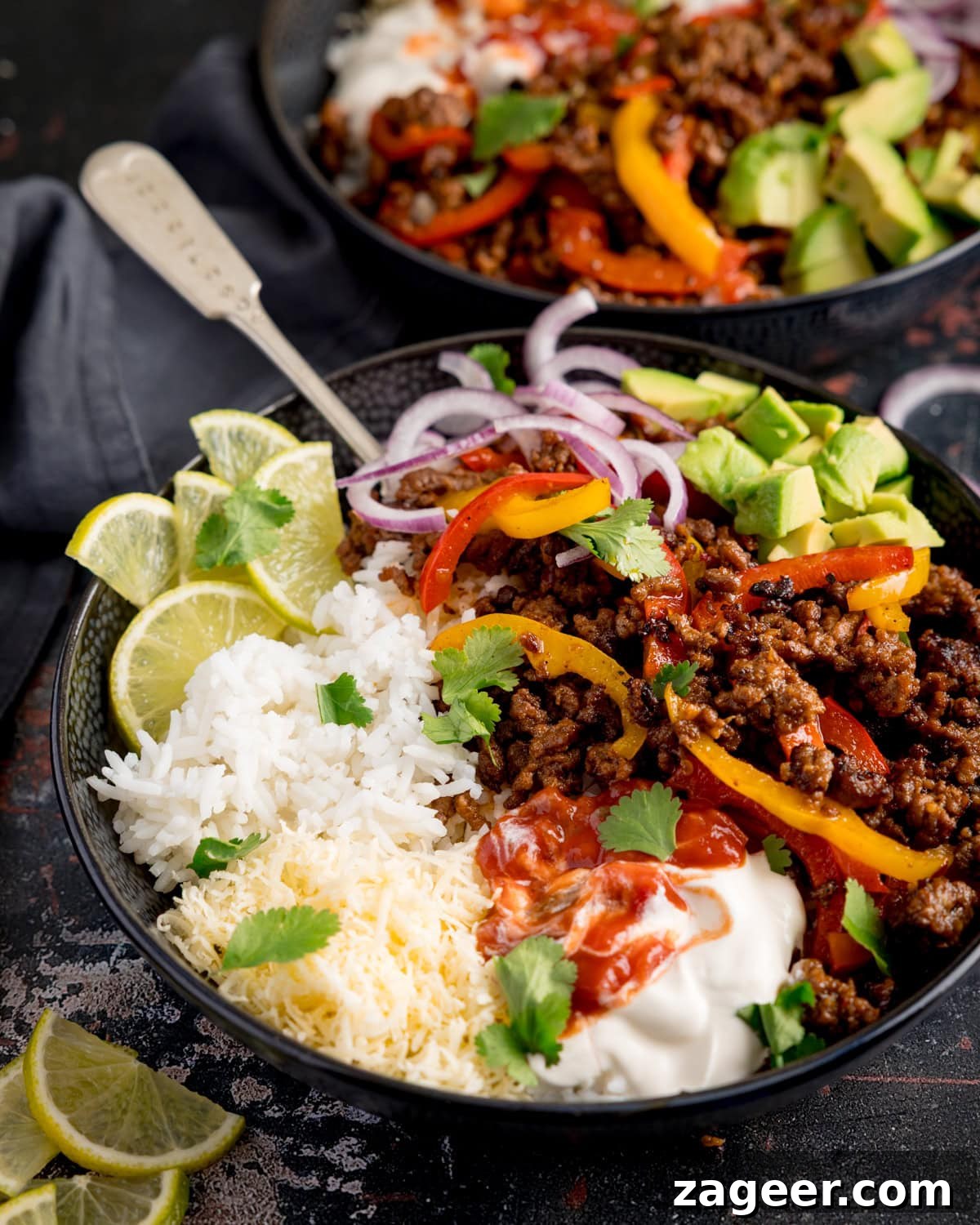 Spiced Minced Beef Fajita Bowls with Peppers and Rice 2 Two minced beef fajita bowls sit on a dark board. Each bowl contains white rice, grated cheese, seasoned minced beef with peppers, sliced red onion, avocado, lime wedges, soured cream, salsa, and coriander.