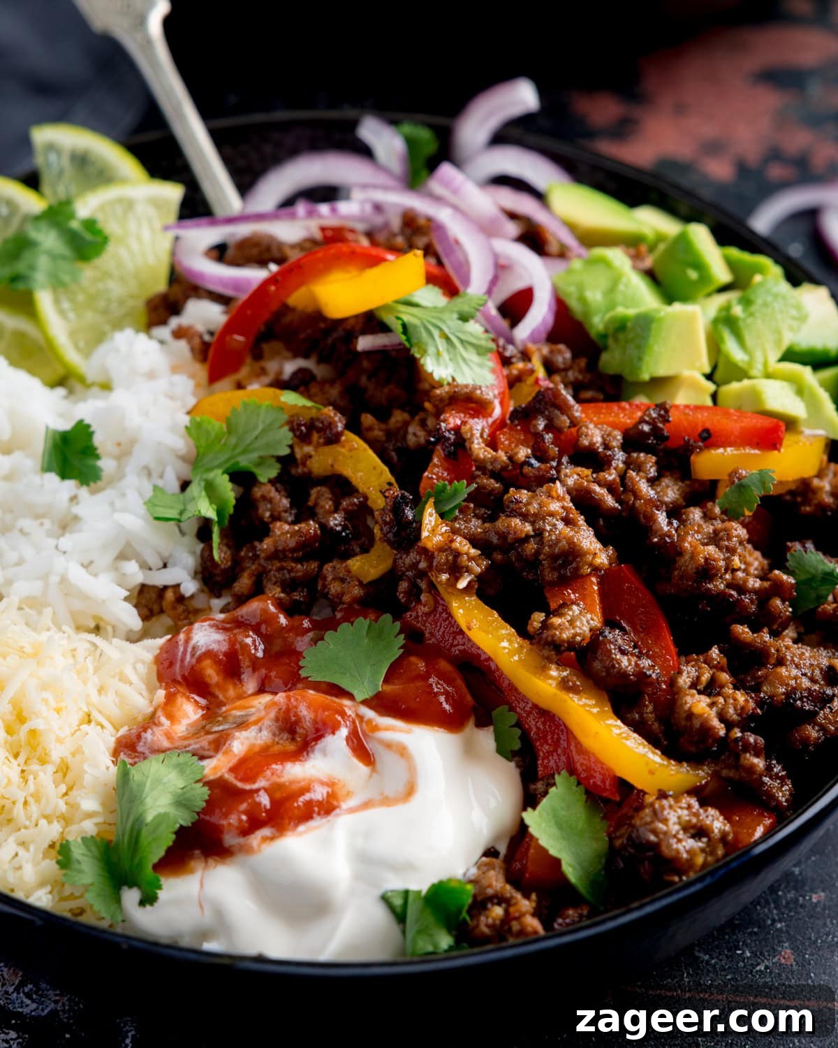 Spiced Minced Beef Fajita Bowls with Peppers and Rice 5 A close up of mince beef fajita bowl filled with seasoned minced beef, sliced peppers, white rice, avocado, grated cheese, soured cream, salsa, red onion, lime wedges, and fresh coriander.