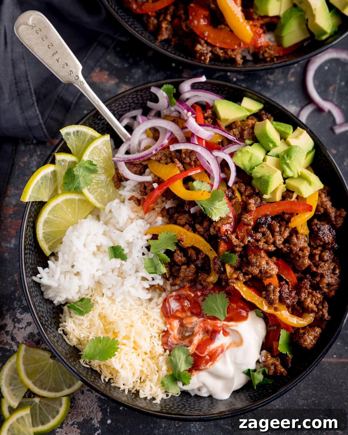 Spiced Minced Beef Fajita Bowls with Peppers and Rice 6 A minced beef fajita bowl with white rice, grated cheese, sliced red onion, peppers, avocado, lime wedges, soured cream, salsa and coriander, served with a spoon.