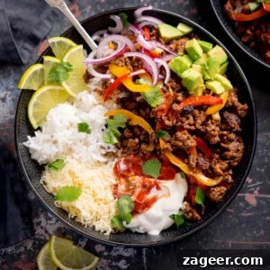A minced beef fajita bowl with white rice, grated cheese, seasoned minced beef with peppers, sliced red onions, avocado, lime wedges, soured cream, salsa, and coriander.
