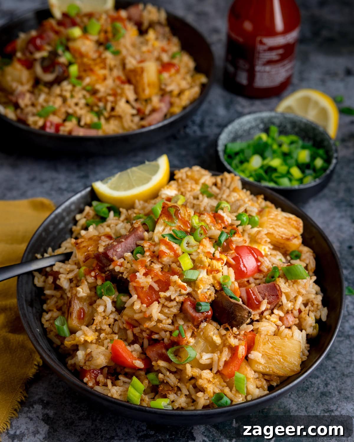 A bowl of ham and pineapple fried rice with chunks of ham, pineapple, pepper and mushroom, topped with chopped spring onions and lemon wedges