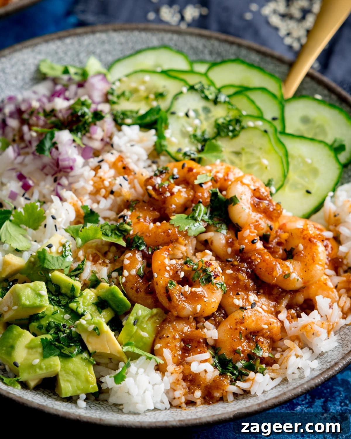 Honey Garlic Prawn Rice Bowl Recipe for Quick Weeknight Meals 2 A close up of a honey garlic prawn rice bowl with; white rice topped with honey glazed prawns, sliced cucumber, diced avocado, chopped red onion, coriander, and sesame seeds.