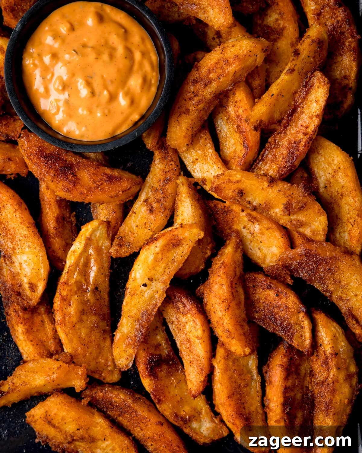 Crispy Oven-Baked Potato Wedges Recipe for Perfectly Seasoned Slices 2 Close up overhead shot of crispy potato wedges on a dark baking tray. A small dish of special sauce for dipping is nestled into the wedges.