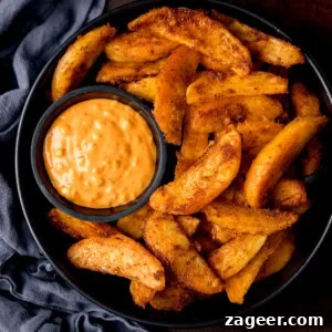 Crispy Oven-Baked Potato Wedges Recipe for Perfectly Seasoned Slices 11 A black bowl filled with crispy potato wedges and a small dish of special sauce for dipping. The bowl is on a dark surface with a blue cloth nearby.