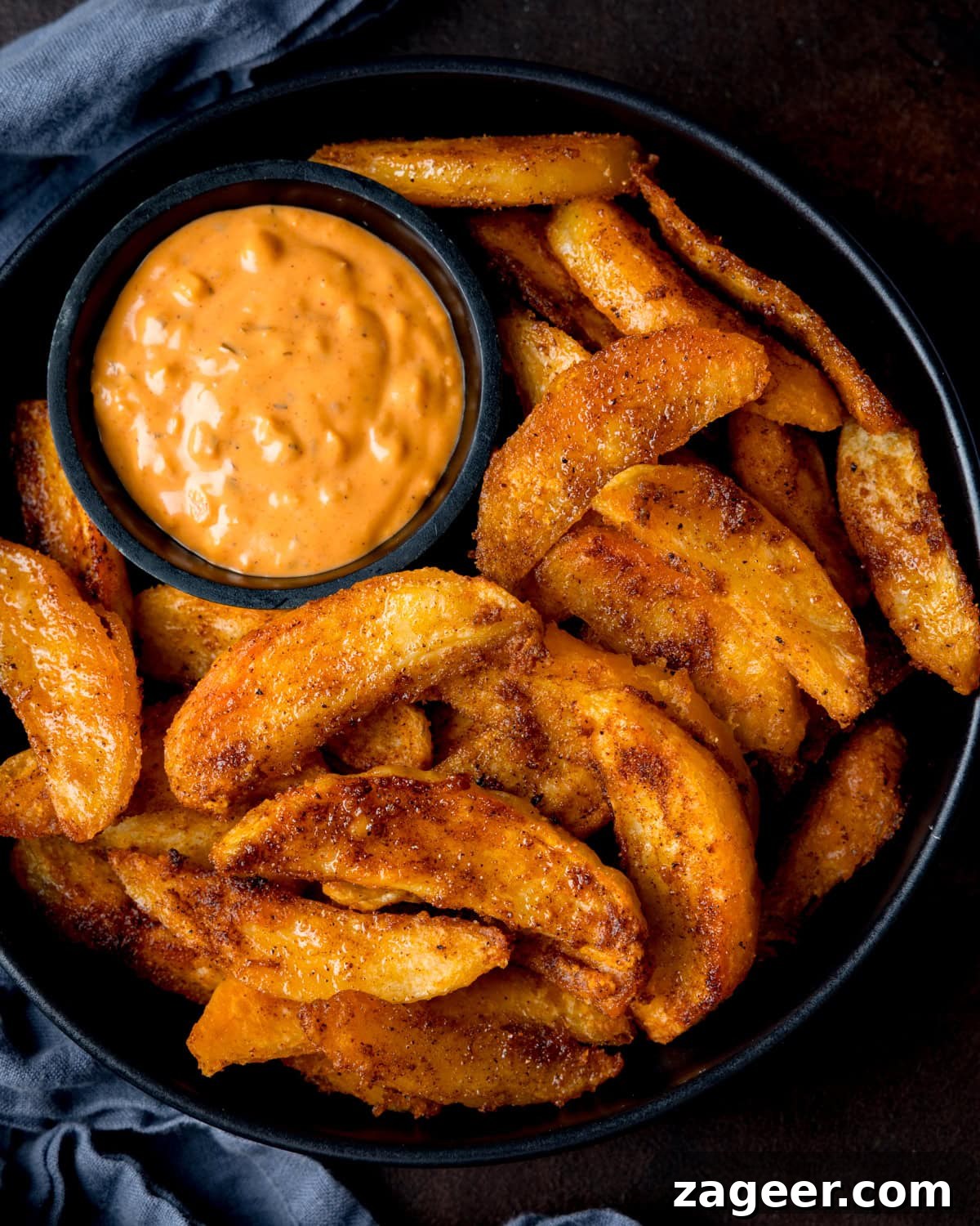 Crispy Oven-Baked Potato Wedges Recipe for Perfectly Seasoned Slices 6 A black bowl filled with crispy potato wedges and a small dish of special sauce for dipping. The bowl is on a dark surface with a blue cloth nearby.
