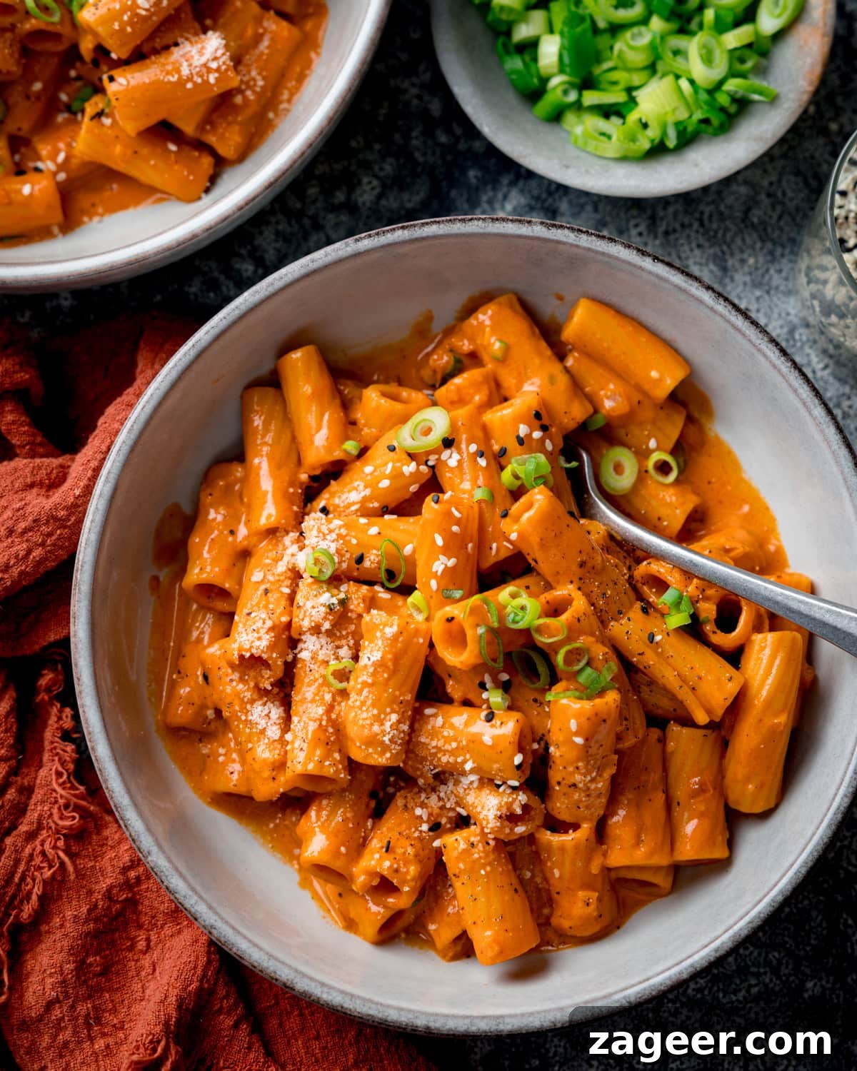 Spicy Gochujang Cream Pasta Recipe You’ll Love 2 A bowl of spicy Gochujang pasta in a creamy gochujang sauce, topped with grated cheese, sliced spring onions, and black pepper, with a fork resting in the bowl. A further bowl of pasta and a dish of spring onions are also in shot.
