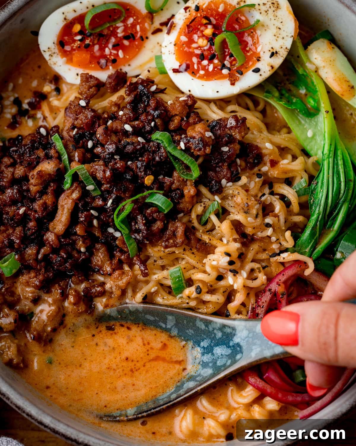 Spicy Pork Curry Ramen Recipe for Bold Flavor 6 Close up of a bowl of Curry Ramen with Spicy Pork, halved soft-boiled eggs, pak choi, pickled onions, spring onions, and rich broth; a hand with red nails is holds a spoon in the bowl to capture the sauce.