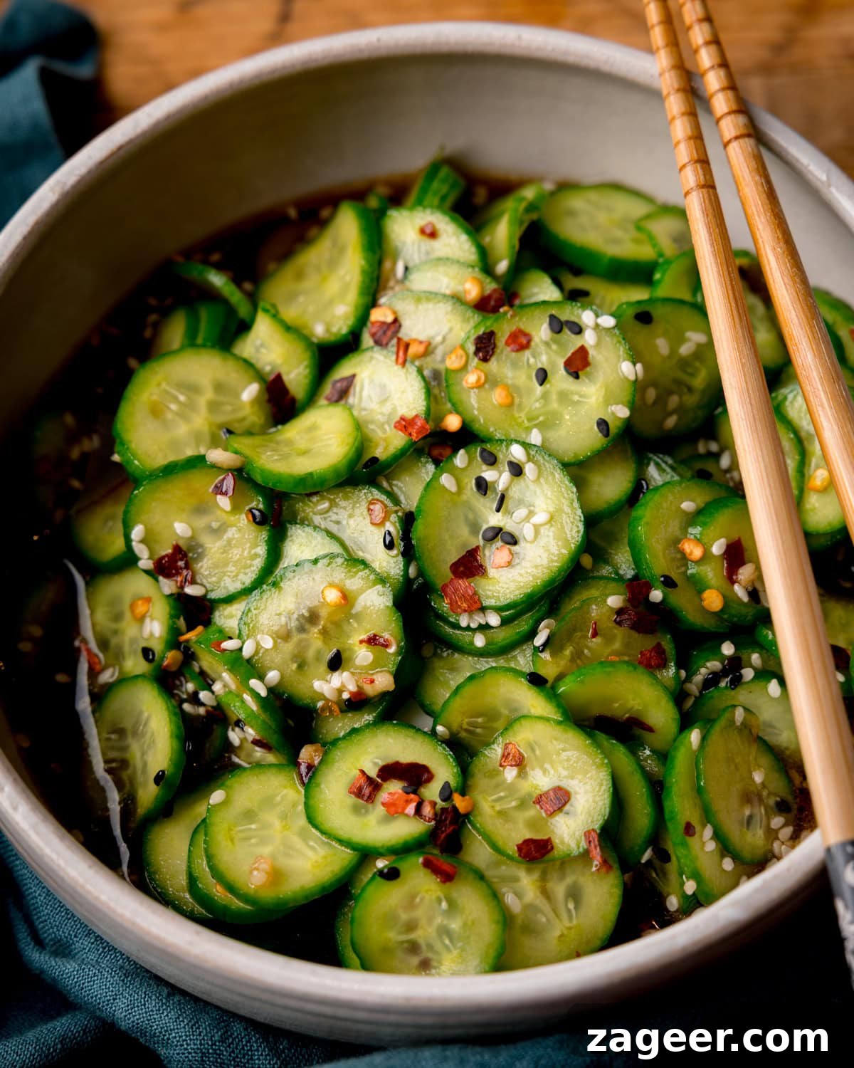 Crisp Asian Cucumber Salad with Sesame Ginger Dressing 2 A bowl of Asian Cucumber Salad, with sliced cucumbers topped with sesame seeds, red chilli flakes, and a soy-sesame sauce, with a pair of chopsticks resting on the bowl.