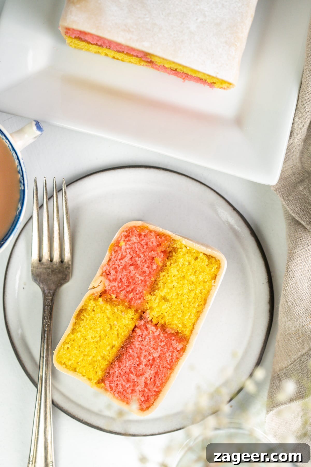 Perfect Battenberg Cake Recipe: Step-by-Step Guide 6 Overhead image of battenberg cake slice on a white plate on a light background. There is a fork on the plate and a cup of tea and more battenberg just in shot.