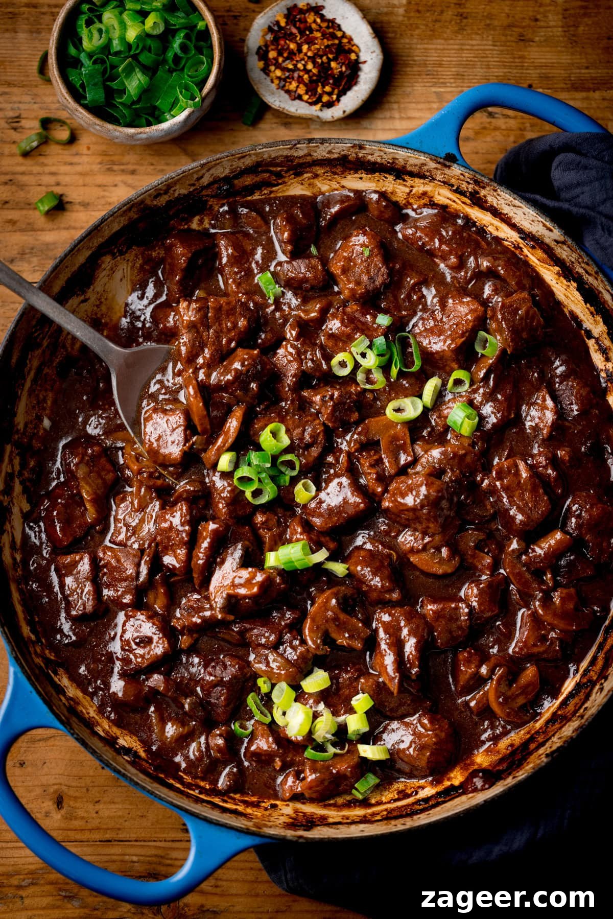 Authentic Chinese-Style Beef Curry Recipe for Home Cooking 2 A blue cast iron casserole filled with Chinese Beef Curry, topped with sliced spring onions. There are little bowls of chopped spring onions and chilli flakes nearby. The pan is on a wooden surface.