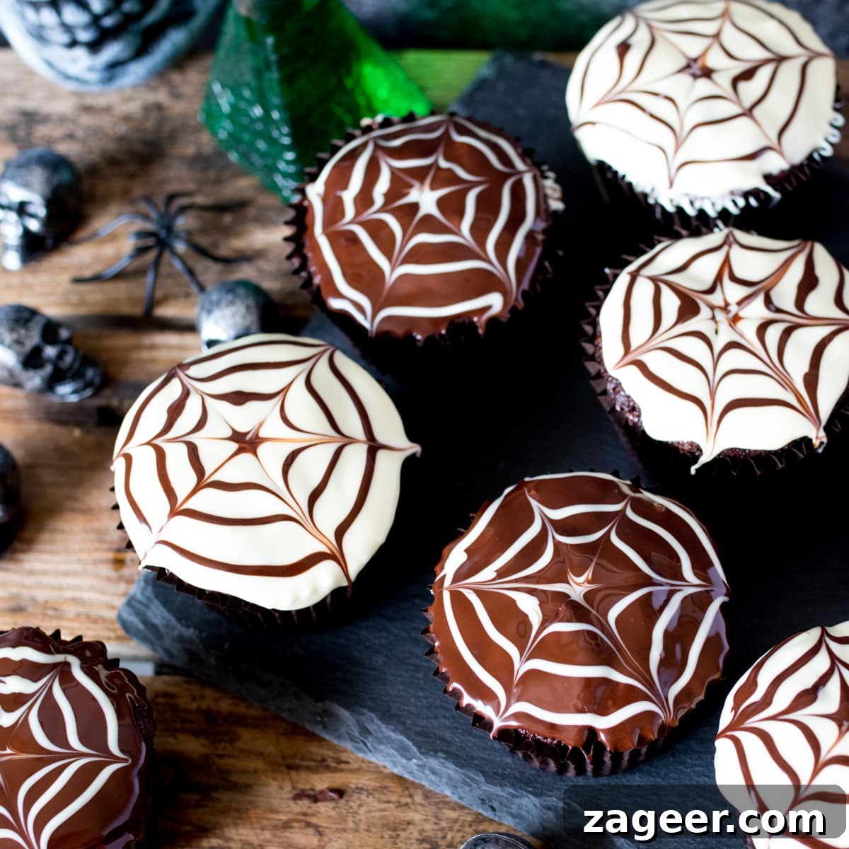 Spider Web Chocolate Cupcakes for Halloween Parties 7 Chocolate Halloween cupcakes with spiderweb patterns in the topping placed on a dark slate background with toy skulls and spiders in the background.