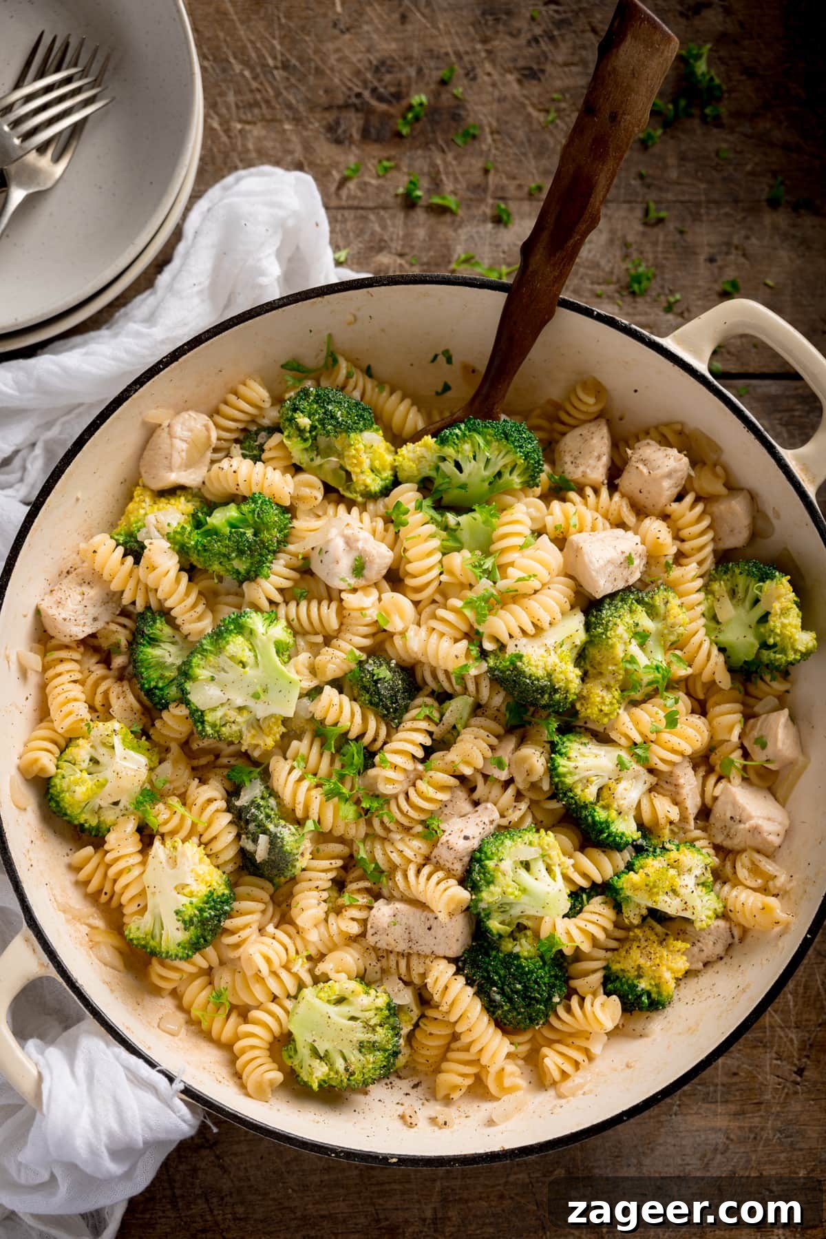 One-Pot Chicken and Broccoli Pasta Dinner in 30 Minutes 2 Overhead shot of chicken and broccoli pasta in a white cast iron pan with a wooden spoon sticking out. The pan is on a wooden table with a white napkin and bowls nearby.