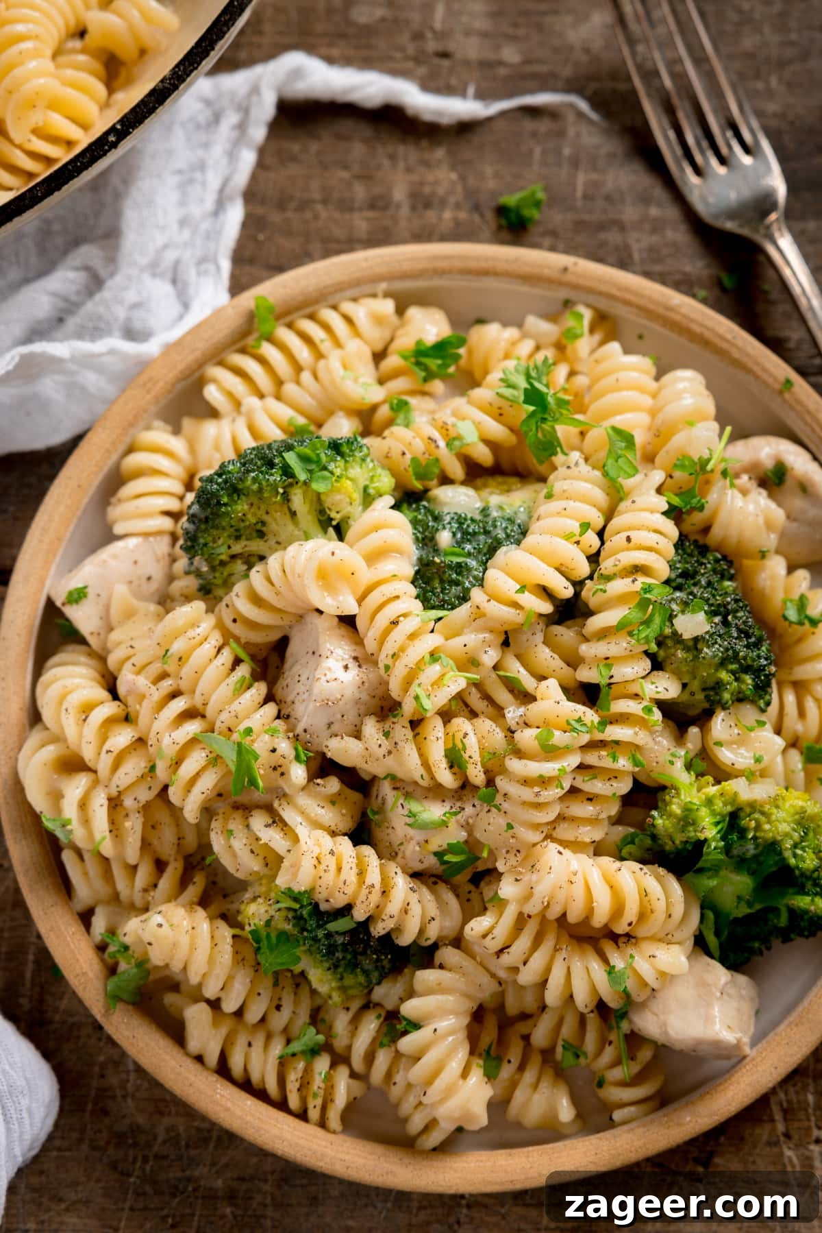 One-Pot Chicken and Broccoli Pasta Dinner in 30 Minutes 5 Overhead shot of chicken and broccoli pasta on a white plate with stone rim. The plate is on a wooden table with a white napkin and fork nearby.