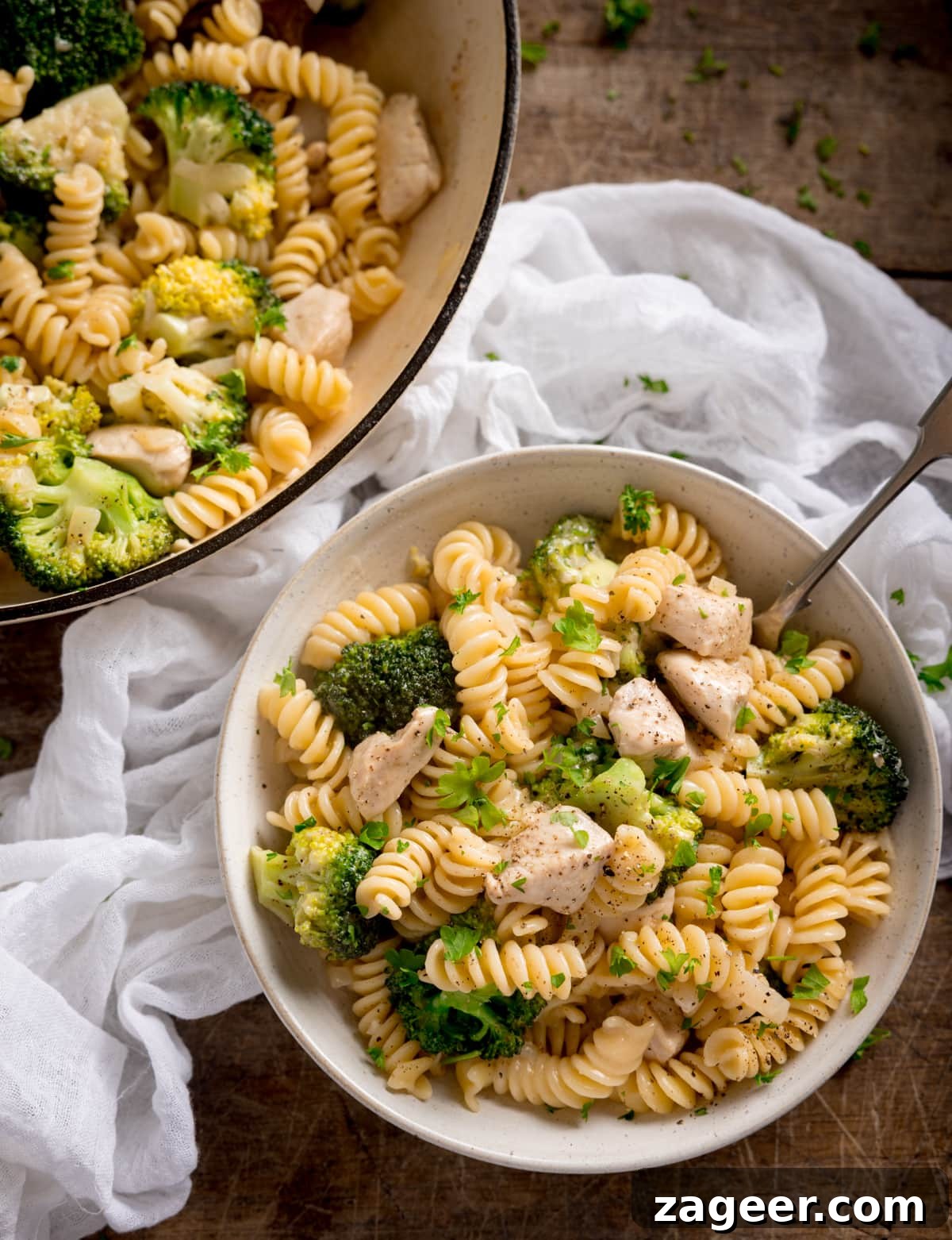 One-Pot Chicken and Broccoli Pasta Dinner in 30 Minutes 6 Overhead shot of chicken and broccoli pasta in a white bowl with a fork sticking out. The bowl is on a wooden table with the pan of pasta and a white napkin nearby.