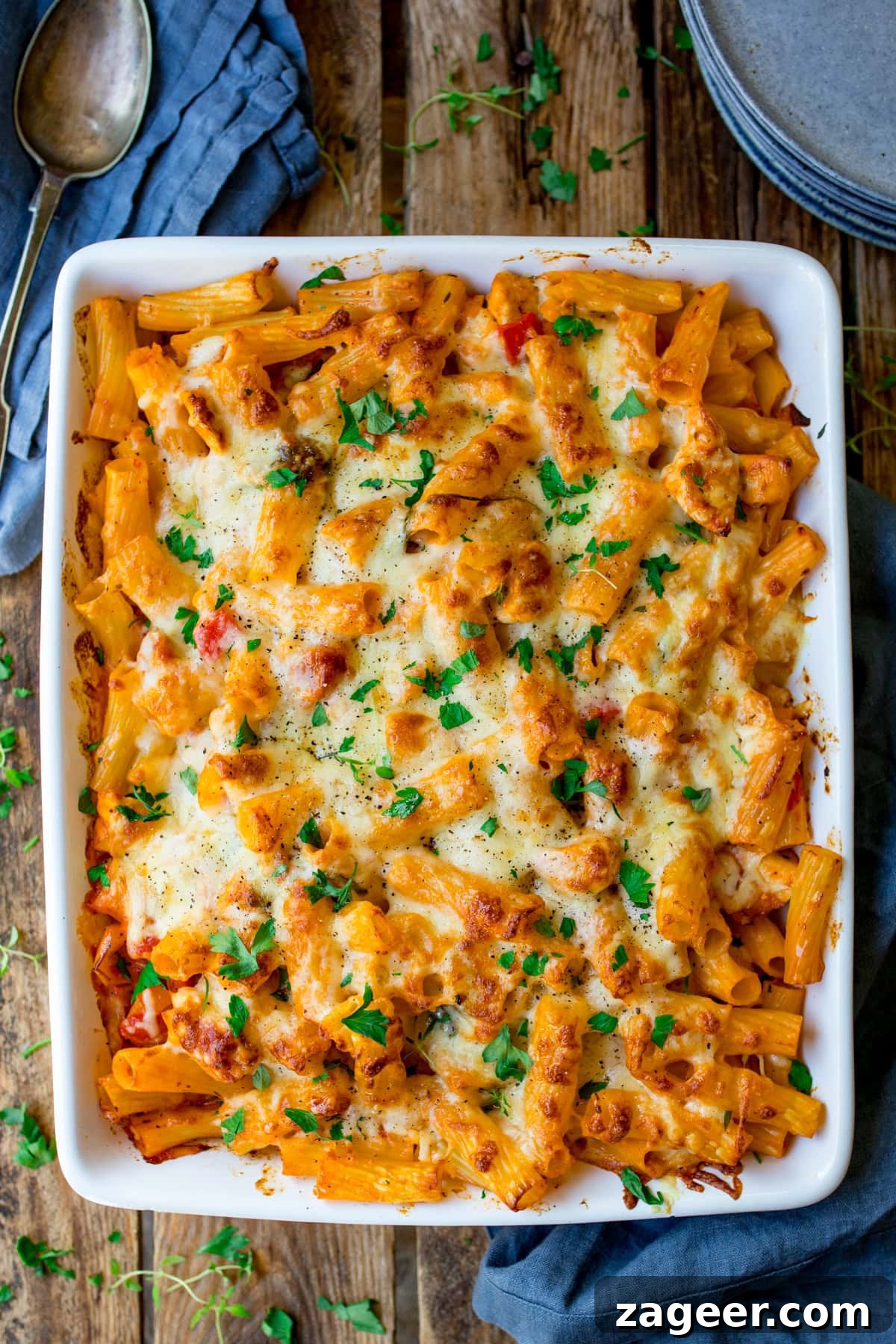 A cheesy chicken bacon pasta bake made with rigatoni, melted cheese, and sprinkled herbs fills a white rectangular dish. The golden brown top is garnished with chopped parsley. This delectable dish rests on a wooden table alongside a blue serviette and spoon.