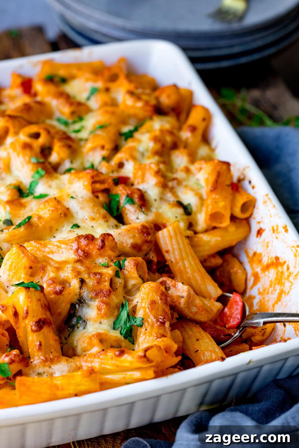 A close-up of a cheesy chicken pasta bake in a white rectangular casserole dish. The pasta is covered in melted cheese and garnished with chopped parsley. A spoon rests in the dish, and the pasta has a golden, crispy texture.