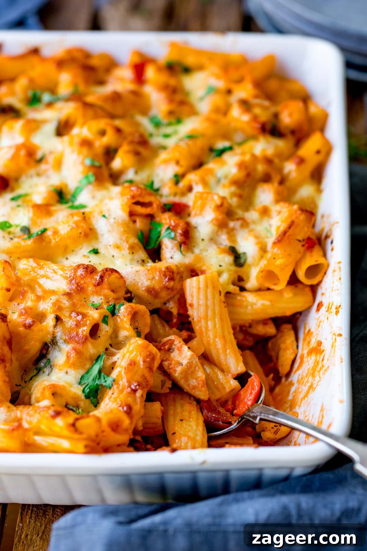 Close-up of a cheesy chicken bacon pasta bake in a white casserole dish. The dish features rigatoni, melted cheese, herbs, and chunks of veg and sauce. A spoon is partially inserted into the savoury blend, while a blue cloth lies nearby.