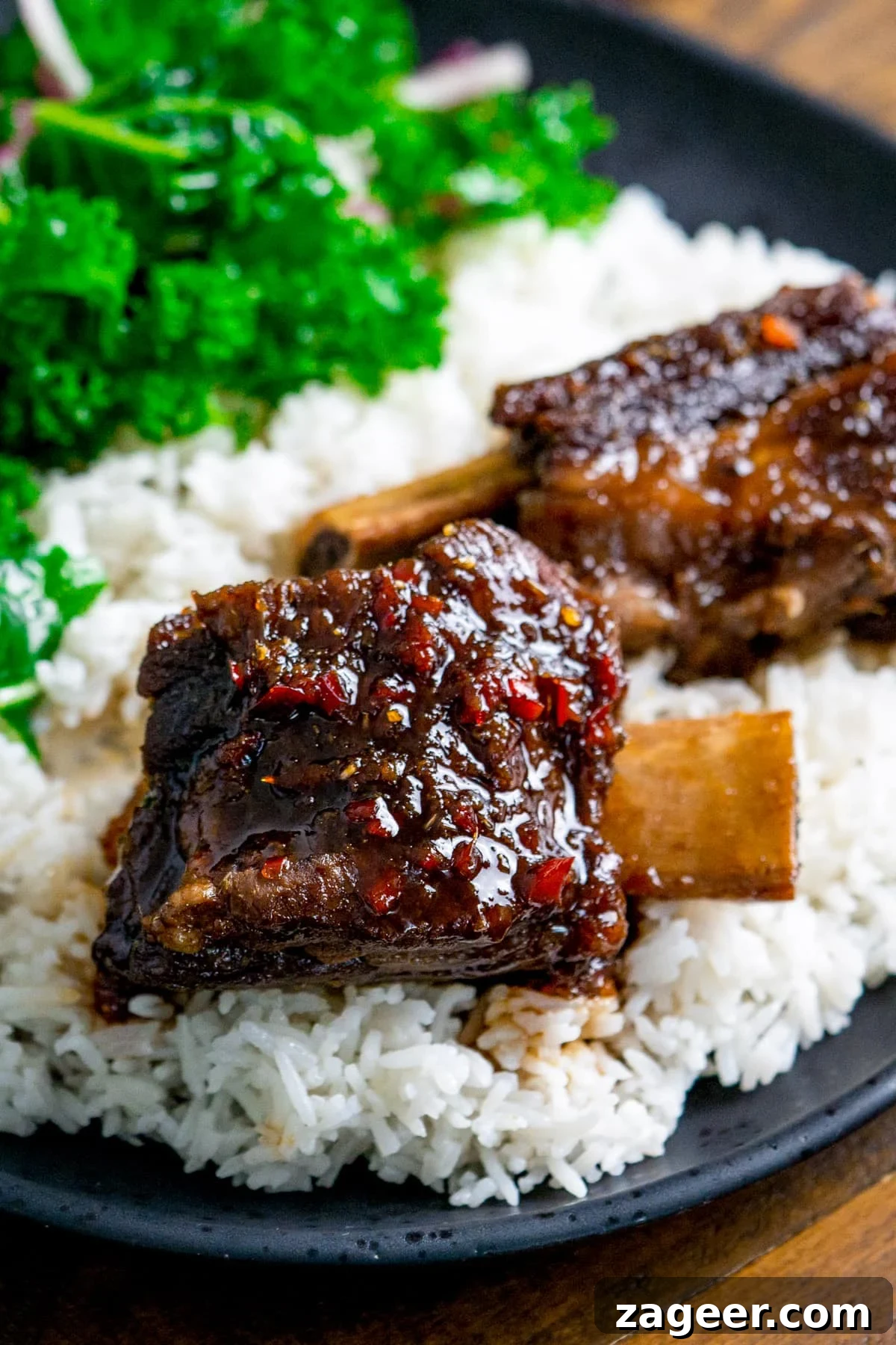Slow cooked short ribs with chilli glaze on a bed of rice on a dark plate. Kale is also on the plate in the background.