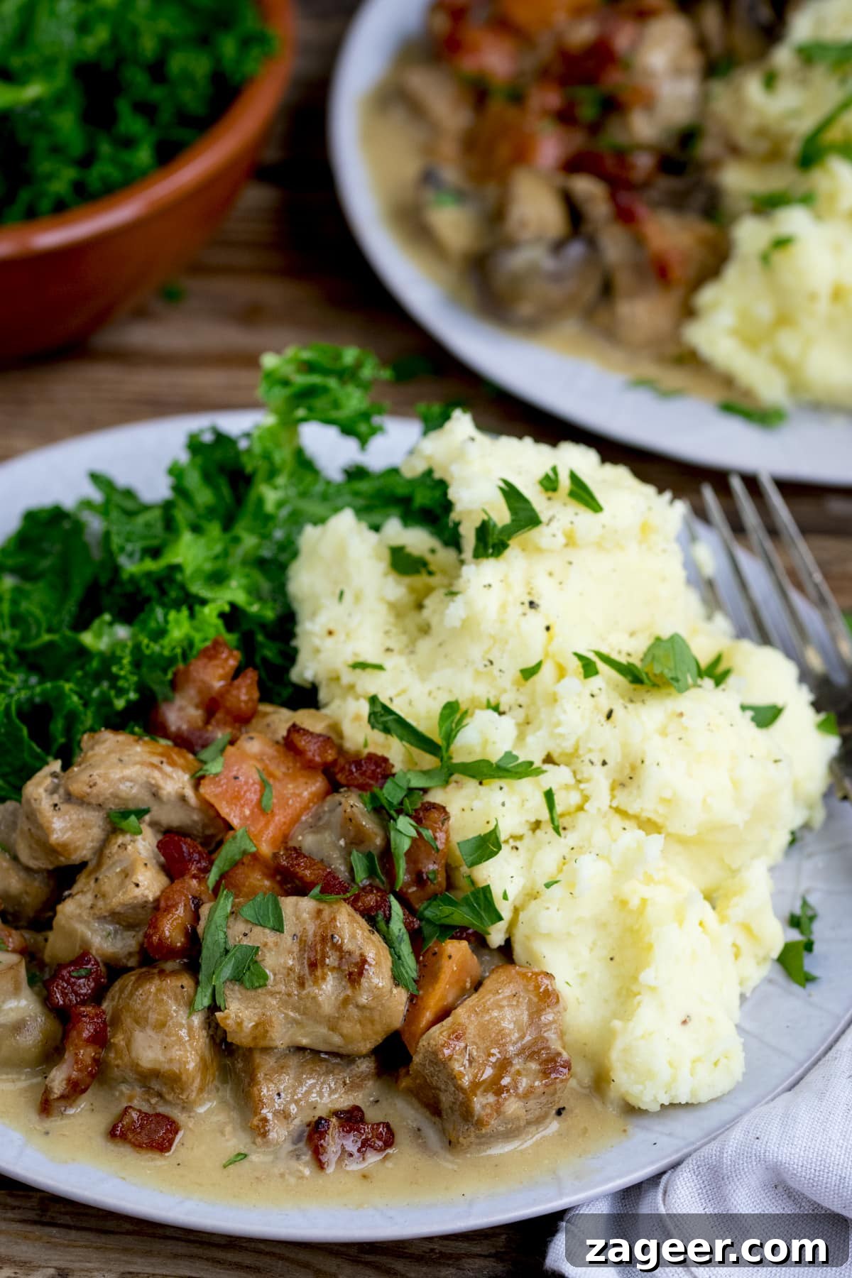 Creamy Slow-Cooked Pork Casserole Recipe for Comforting Dinners 2 Creamy pork casserole, topped with bacon on a plate with mashed potato and kale. There is a further plate and a bowl of kale in the background.