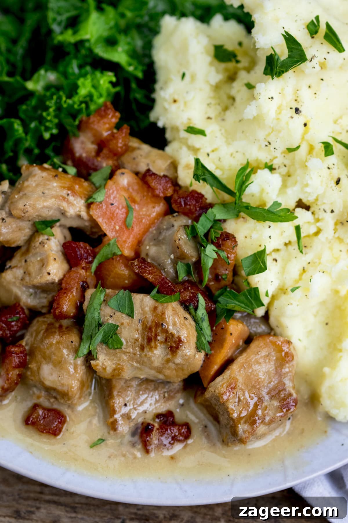Creamy Slow-Cooked Pork Casserole Recipe for Comforting Dinners 6 Close-up overhead image of cream pork casserole with bacon, carrot and mushroom on a light plate with mashed potato and kale. Parsley has been sprinkled on top.