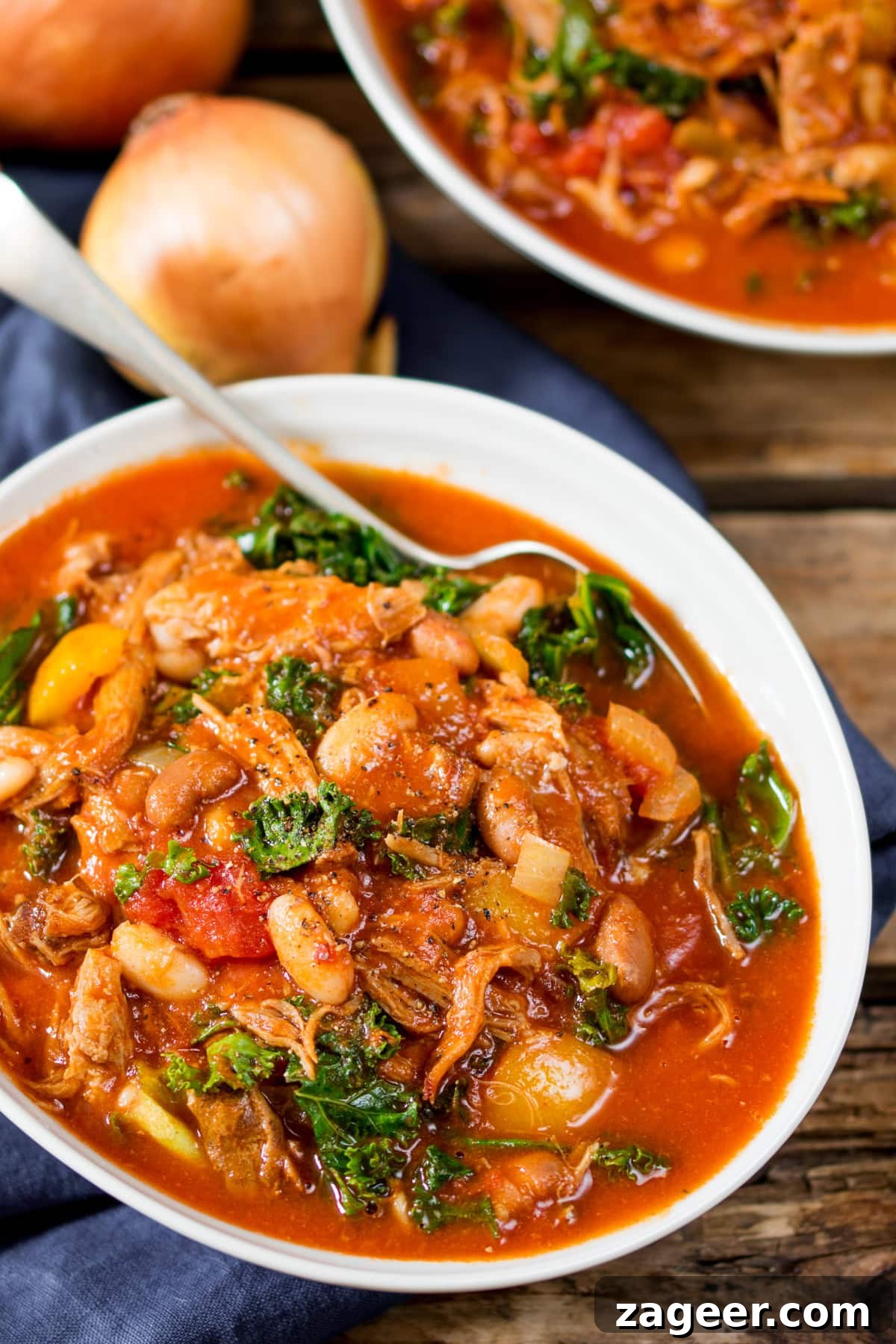 Hearty Pulled Pork and White Bean Soup Recipe 2 A white bowl of hearty pulled pork and bean soup with kale and vegetables, served in a rich tomato-based stew, with a spoon and onions in the background.