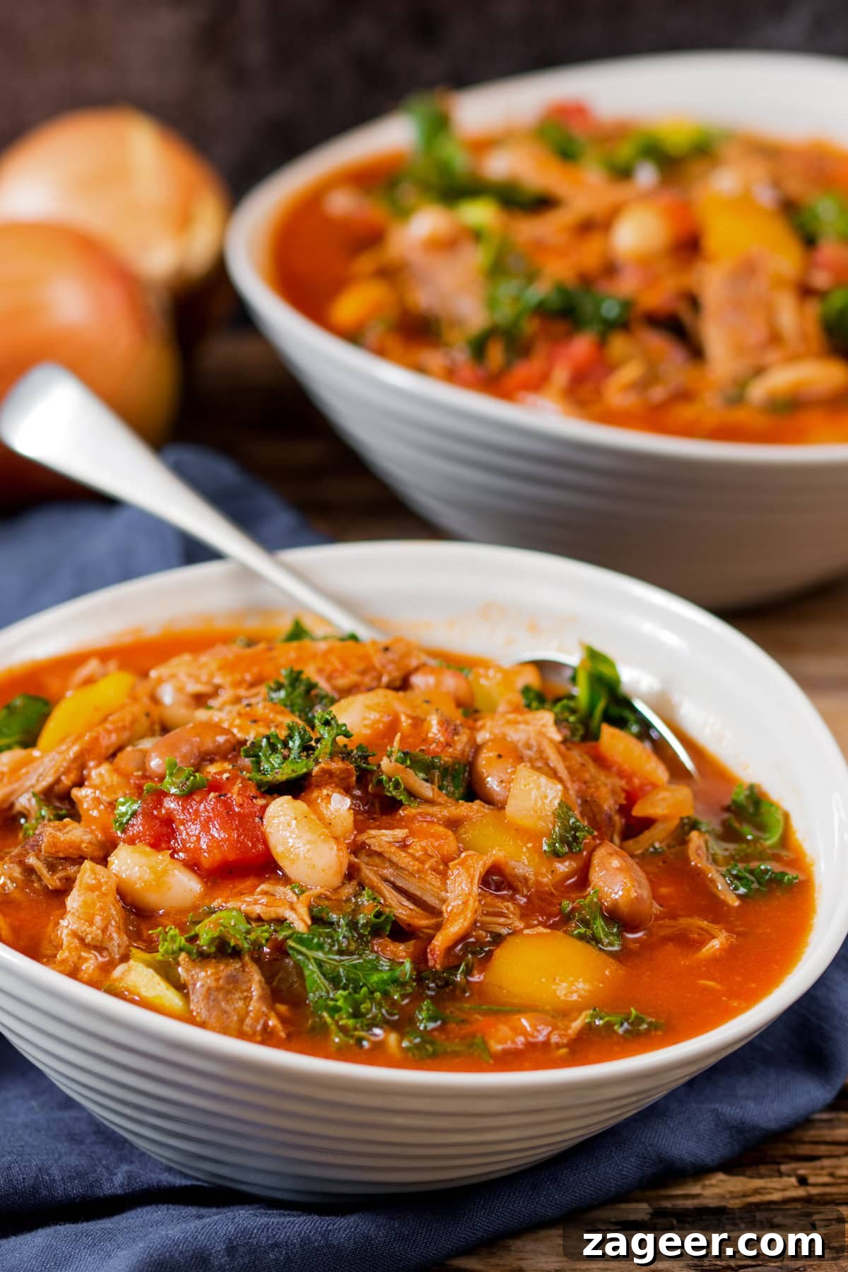 Hearty Pulled Pork and White Bean Soup Recipe 5 Two white bowls filled with hearty pulled pork and bean soup, featuring shredded meat, kale, tomatoes, and stock, with a spoon resting in the front bowl and 2 onions in the background slightly out of focus.