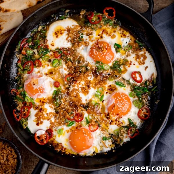 Spicy Scrambled Eggs with Smashed Beans Breakfast Bowl 7 Square image of four sunny-side-up fried eggs in a skillet, topped with natural yogurt, fried onions, sliced red chiles, scallions, and sesame seeds. Some flatbreads are in the corner of the image.