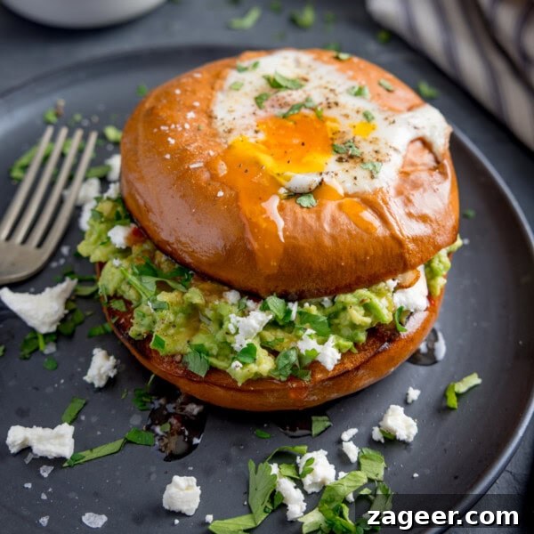 Spicy Scrambled Eggs with Smashed Beans Breakfast Bowl 8 A bagel with a fried egg, smashed avocado, crumbled cheese, and chopped herbs sits on a dark plate with a fork beside it.
