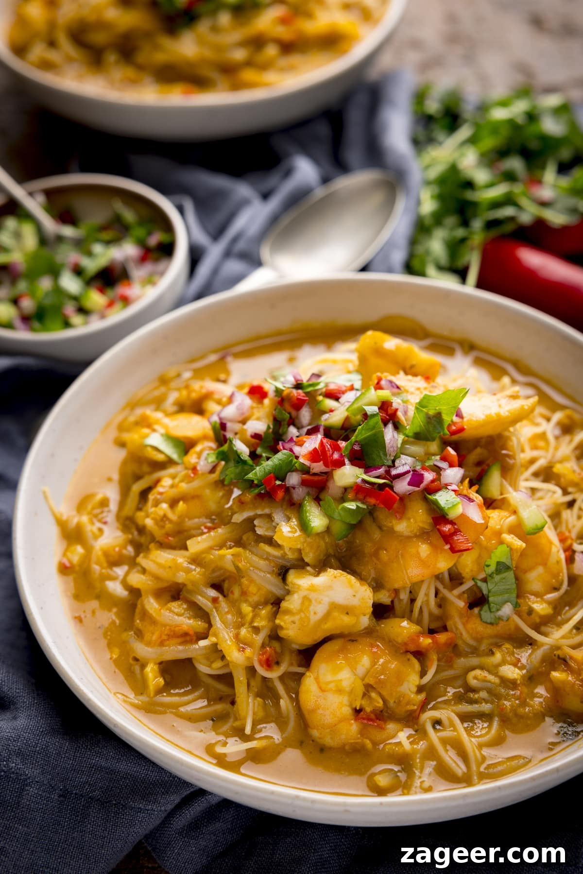 Creamy Seafood Laksa Recipe: Authentic Malaysian Coconut Noodle Soup 2 Seafood laksa in a white bowl, on a wooden table. There is a napkin, spoon and ingredients scattered around.