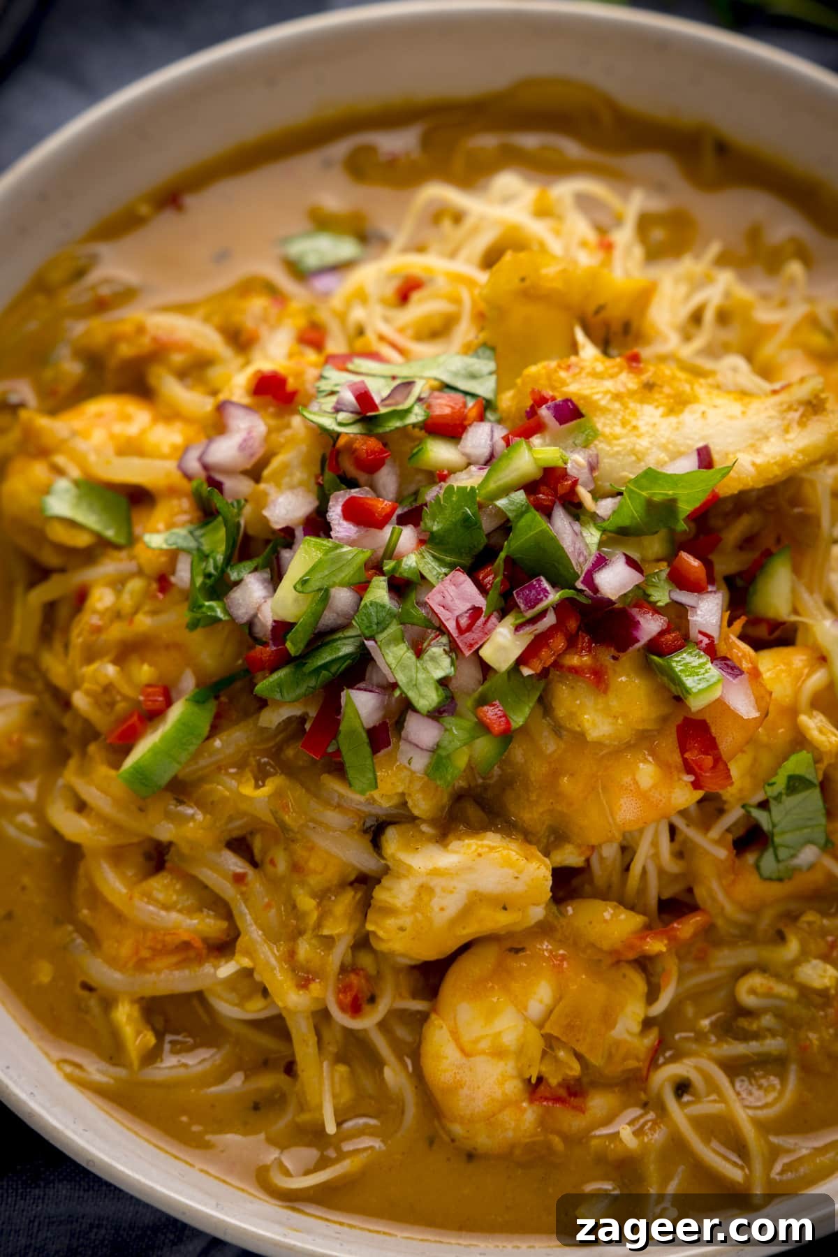 Creamy Seafood Laksa Recipe: Authentic Malaysian Coconut Noodle Soup 5 Close up of seafood laksa in a white bowl topped with a chilli-coriander and red onion garnish.