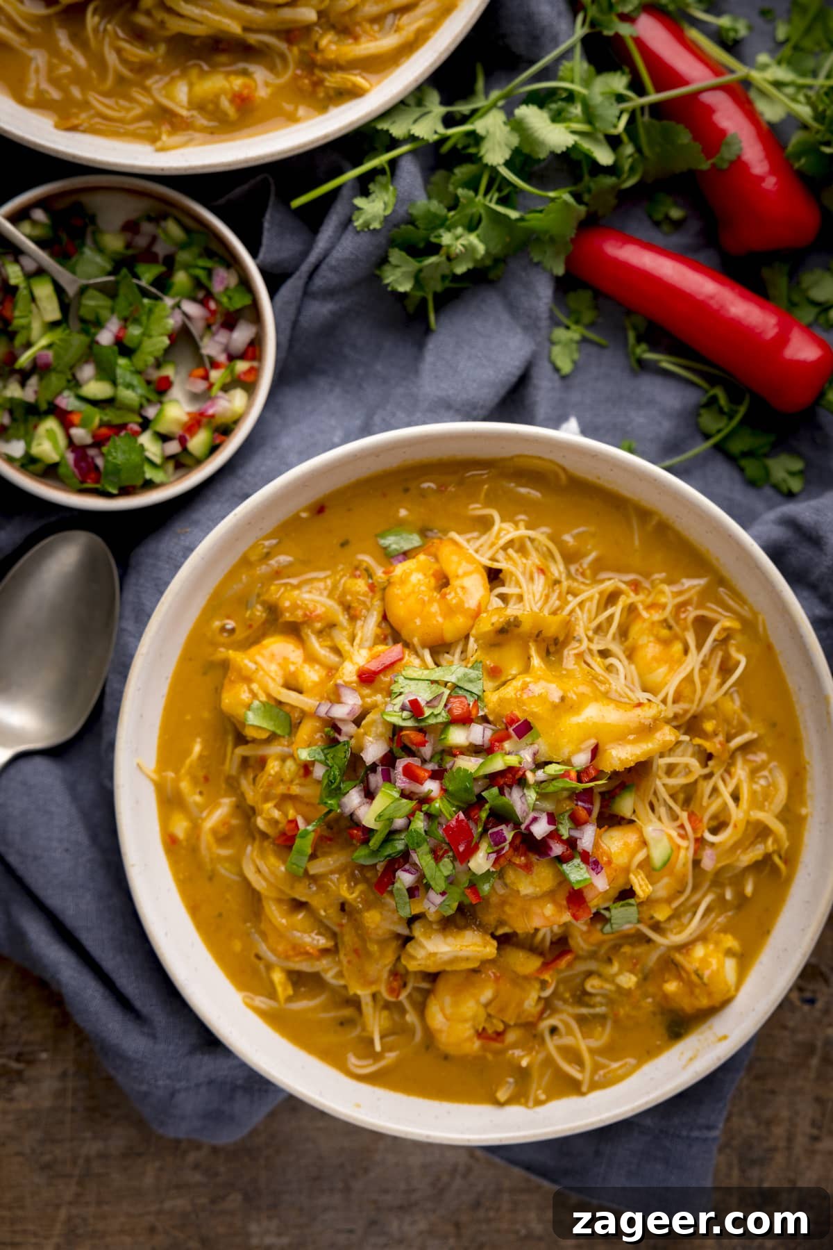 Creamy Seafood Laksa Recipe: Authentic Malaysian Coconut Noodle Soup 6 Overhead image of seafood laksa in a white bowl on a wooden table. There is a spoon, blue napkin, a garnish bowl, a second bowl of laksa and some chillies and coriander around the main bowl.