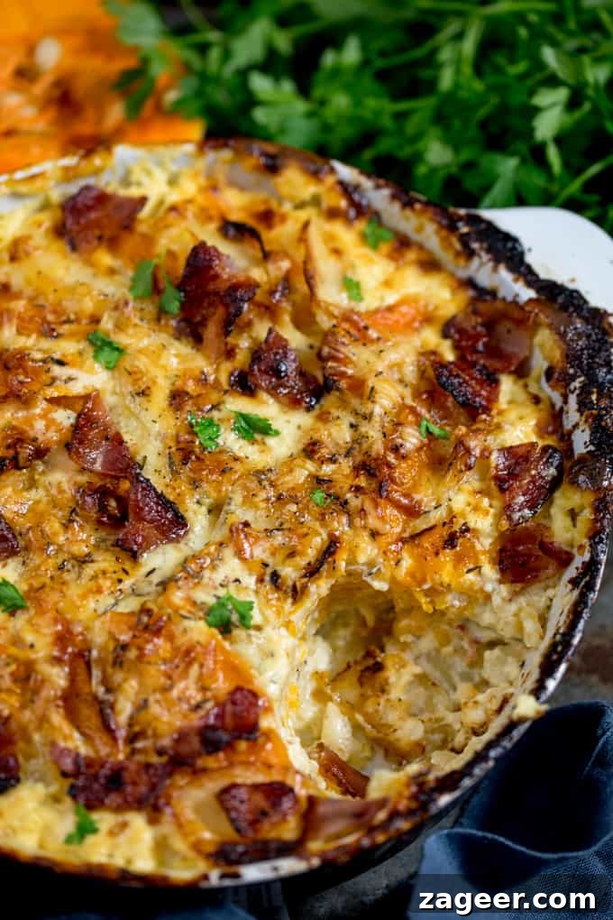 Close up of Squash and Potato Gratin in a grey and white baking dish with a scoop taken out, topped with bacon and parsley in the background
