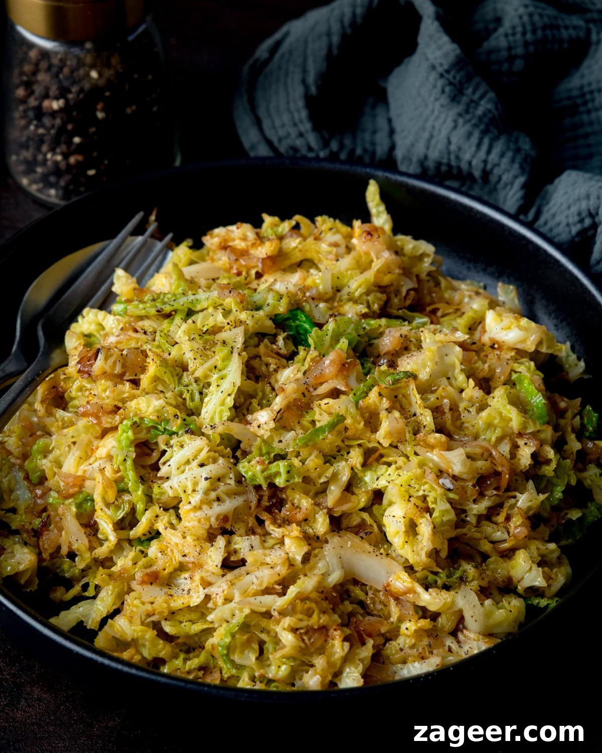 Pan Sautéed Cabbage with Garlic and Crispy Edges 2 Side-on image of a black bowl filled with sautéed cabbage, next to a fork, knife, peppermill and dark napkin.