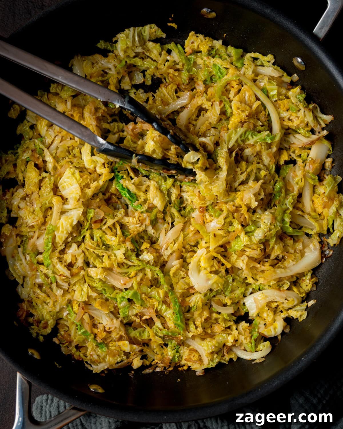 Pan Sautéed Cabbage with Garlic and Crispy Edges 5 Overhead image of a pan filled with sautéed cabbage and onions, with metal tongs resting on top.
