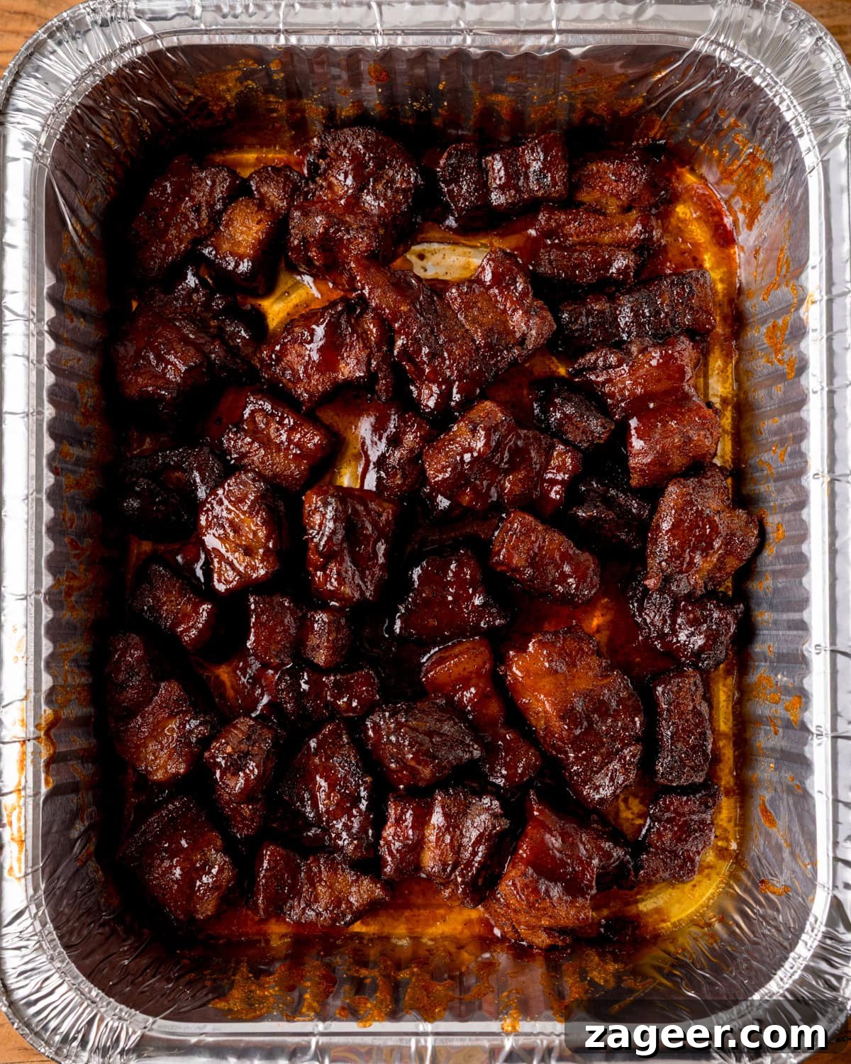 Cubed pieces of cooked, glazed pork belly arranged in a disposable foil tray, each coated with a rich caramelized sauce - Pork Belly Burnt Ends.