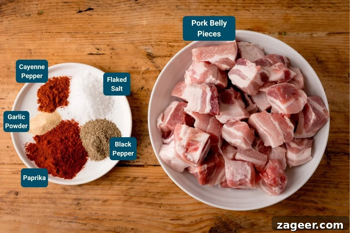 A bowl of raw pork belly pieces sits beside a small plate with paprika, garlic powder, cayenne pepper, black pepper, and flaked sea salt on a wooden surface.