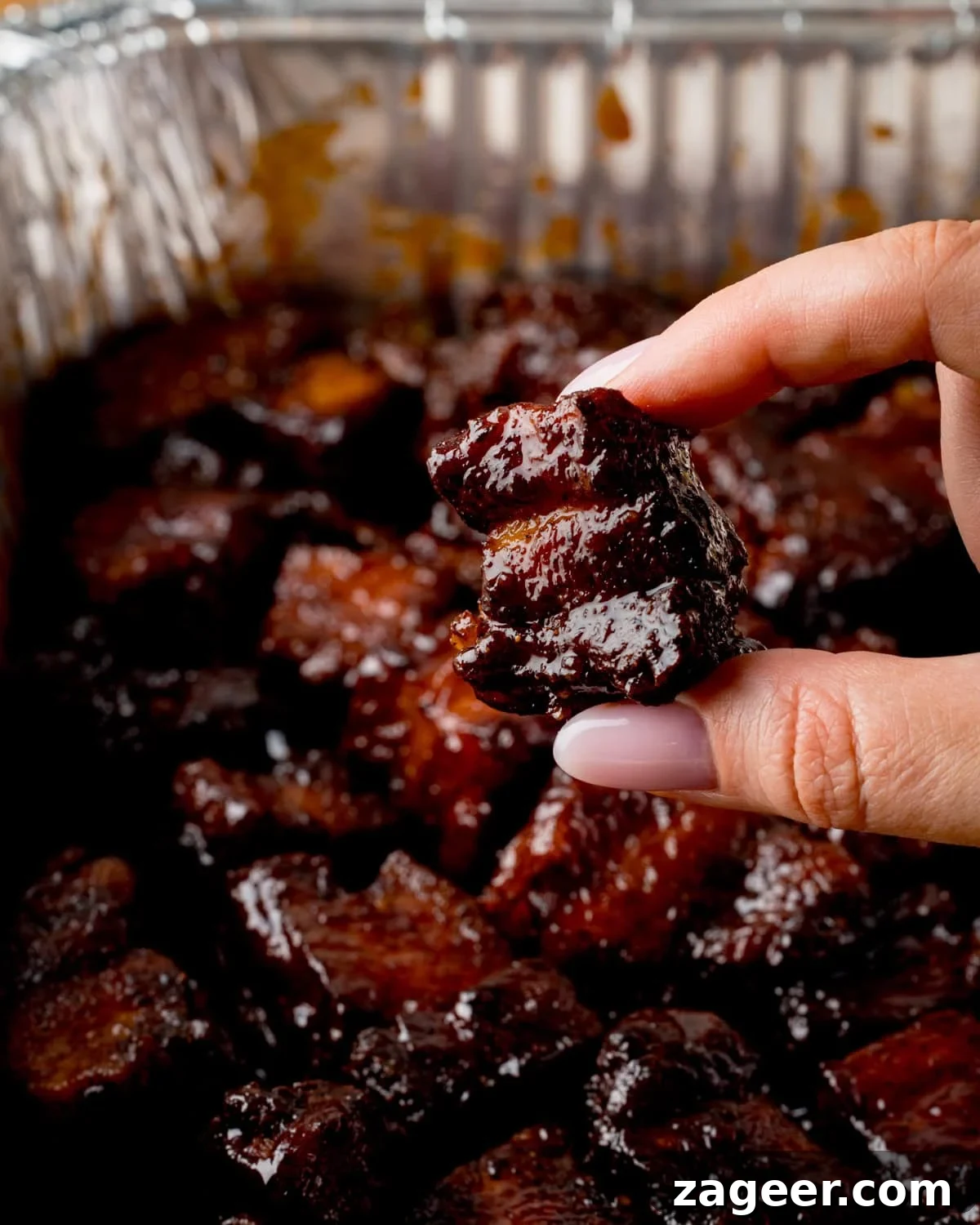 A hand has picked up and is squeezing a a piece of glazed Pork Belly Burnt End - above a tray filled more pork belly bites.