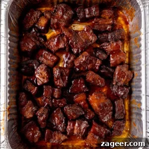 Overhead image of chunks of glazed Pork Belly Burnt Ends arranged in a single layer inside a disposable aluminium tray, coated in a dark, shiny sauce.