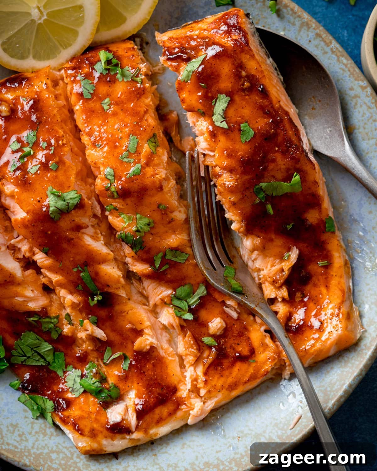 Glazed side of salmon sliced into fillets and garnished with chopped parsley on a light blue plate, with a fork flaking one fillet and lemon slices on the side.