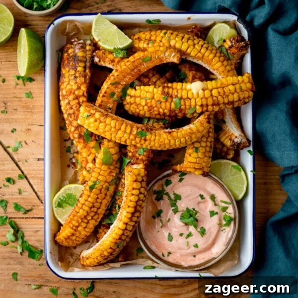 Overhead image of corn ribs served in a tray with lime wedges, chopped herbs, and a bowl of creamy dipping sauce on a wooden table.