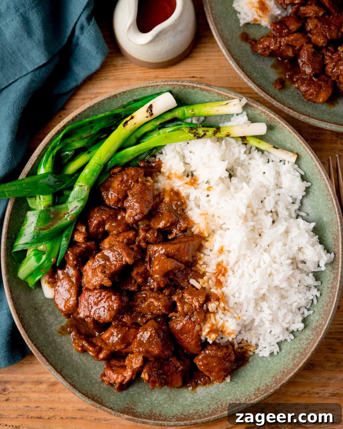 A green plate with white rice, Vietnamese-style caramel pork and grilled spring onions (scallions), with a fork on the side. The plate is on a wooden surface. There is a further plate of pork and a small jug of sauce nearby.