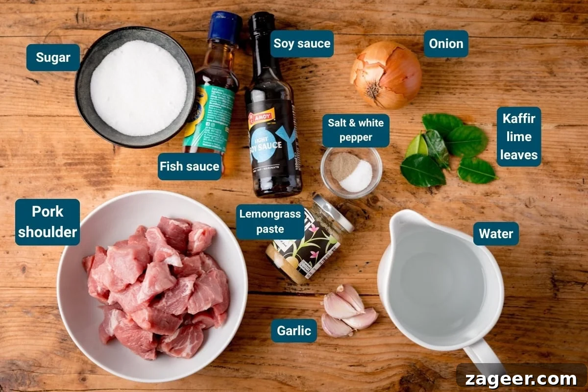 Ingredients for Caramel Pork on a wooden table: pork shoulder, garlic, onion, kaffir lime leaves, lemongrass paste, sugar, fish sauce, soy sauce, pepper and water. All ingredients are labelled.