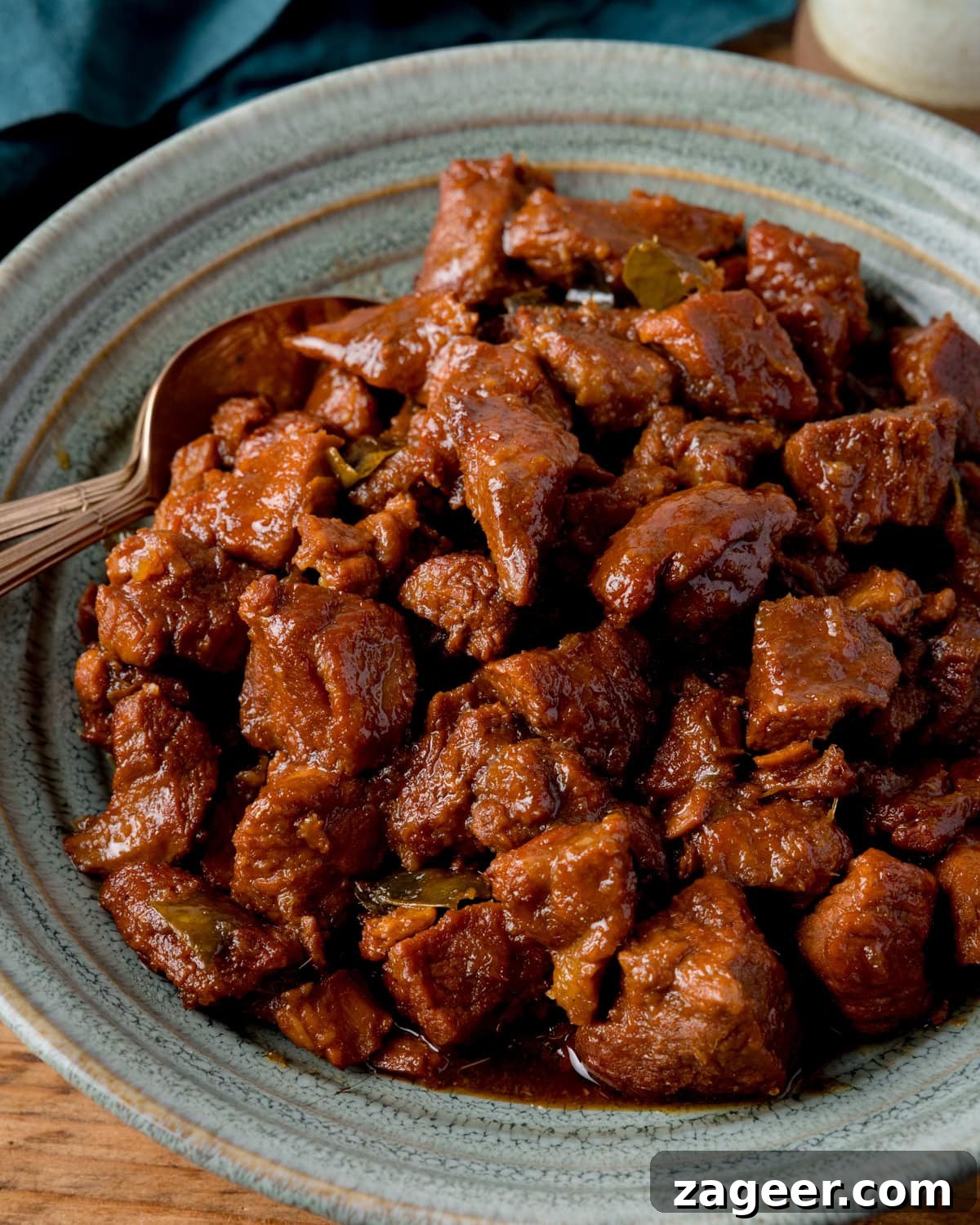 A grey-green bowl filled with saucy, cooked chunks of caramel pork sits a wooden table with two spoons nestled in the pork.