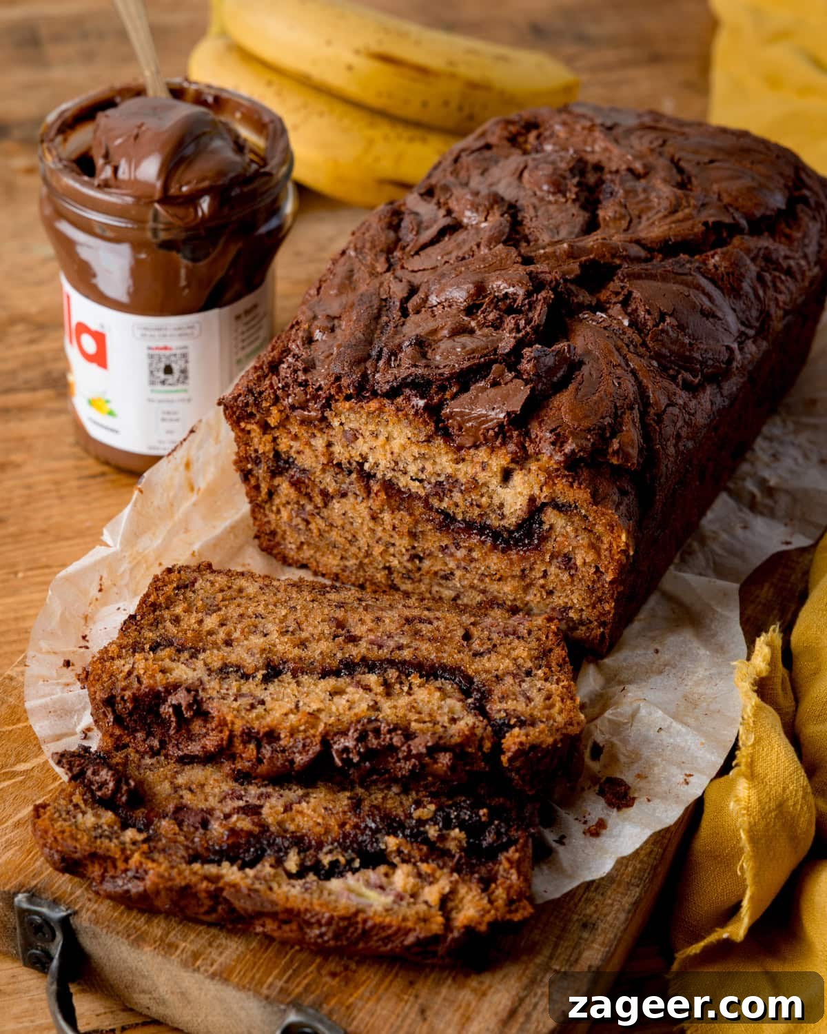A loaf of banana bread with chocolate swirls is sliced on baking parchment next to a jar of Nutella and bananas on a wooden table.