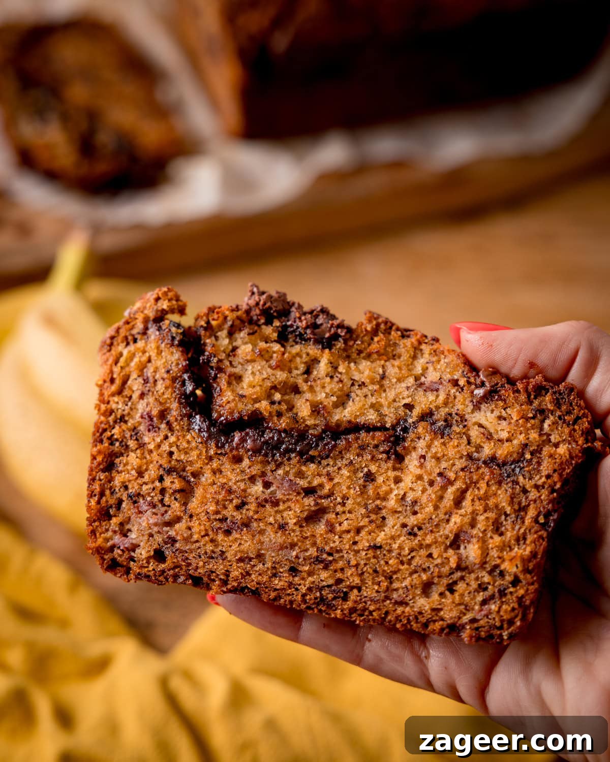 A hand holds a slice of Nutella Banana Bread with a chocolate swirl, with the remaining loaf and a banana in the background on a wooden surface.