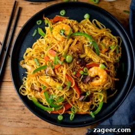 A black plate of stir-fried Singapore noodles with prawns, chicken, car siu pork vegetables, and spring onions, on a wooden table with chopsticks beside it.